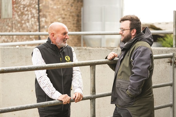 Two people standing by metal fencing in a farm pen, talking with buildings and equipment in the background.