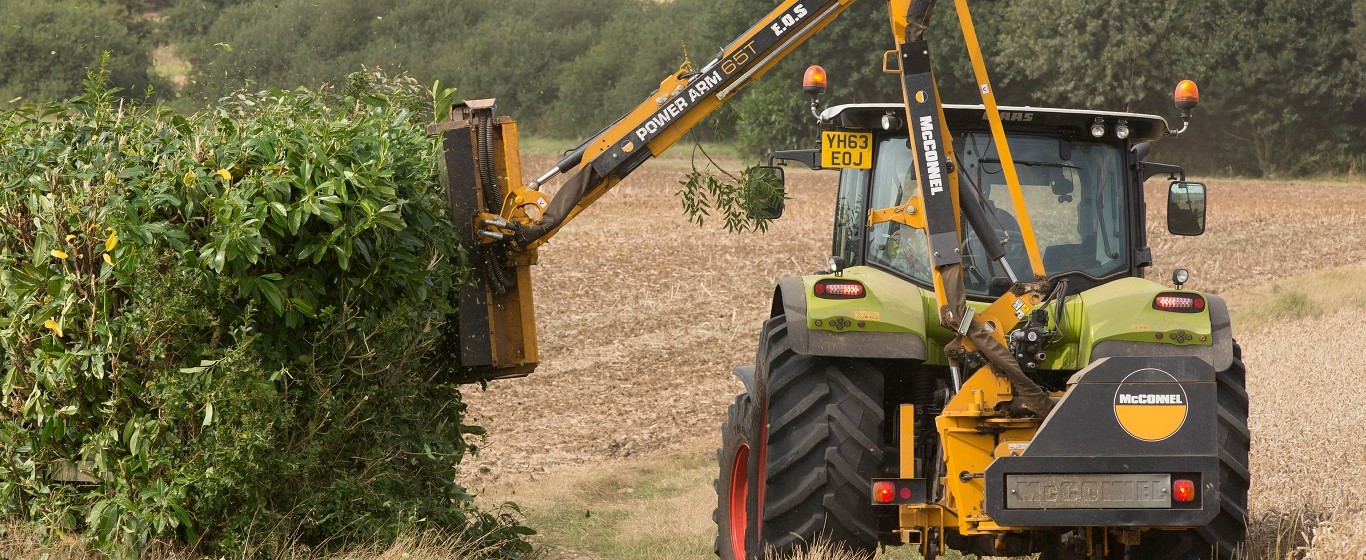 Tractor fitted with a hedge-cutting attachment trimming a roadside hedgerow beside a harvested field in a rural landscape.