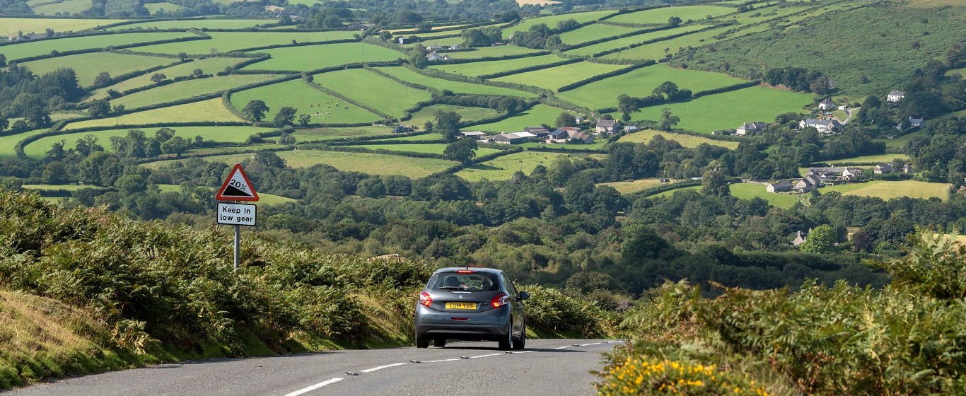Car driving along a rural road with a steep‑hill warning sign, overlooking patchwork green fields and scattered homes in the valley.