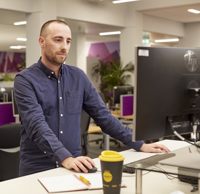 Man working at a standing desk in a modern office, using a computer with a notebook and takeaway coffee cup on the desk.