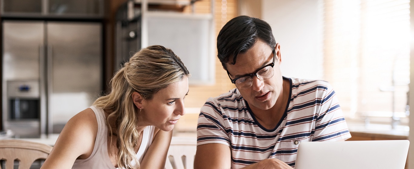 Couple sitting at a kitchen table reviewing information together on a laptop, focused and discussing details in a bright home setting.