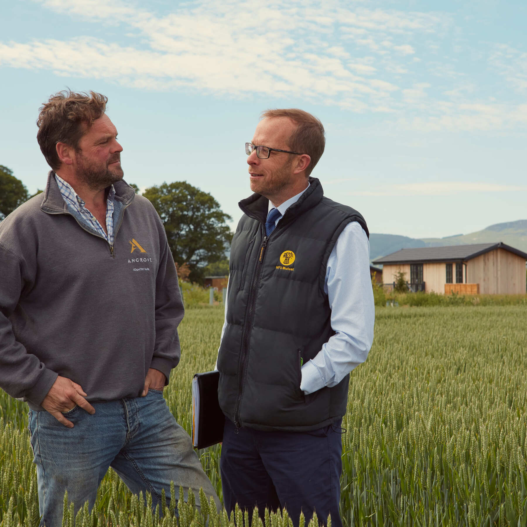 Two men talking in a wheat field, with rural homes and hills in the background.