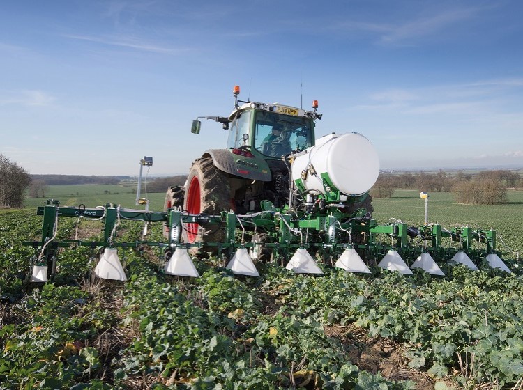 A tractor with mounted agricultural equipment moves through a crop field on a clear day.