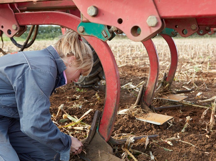 A person in overalls kneels beneath a large piece of red agricultural machinery, inspecting or adjusting parts above the soil.