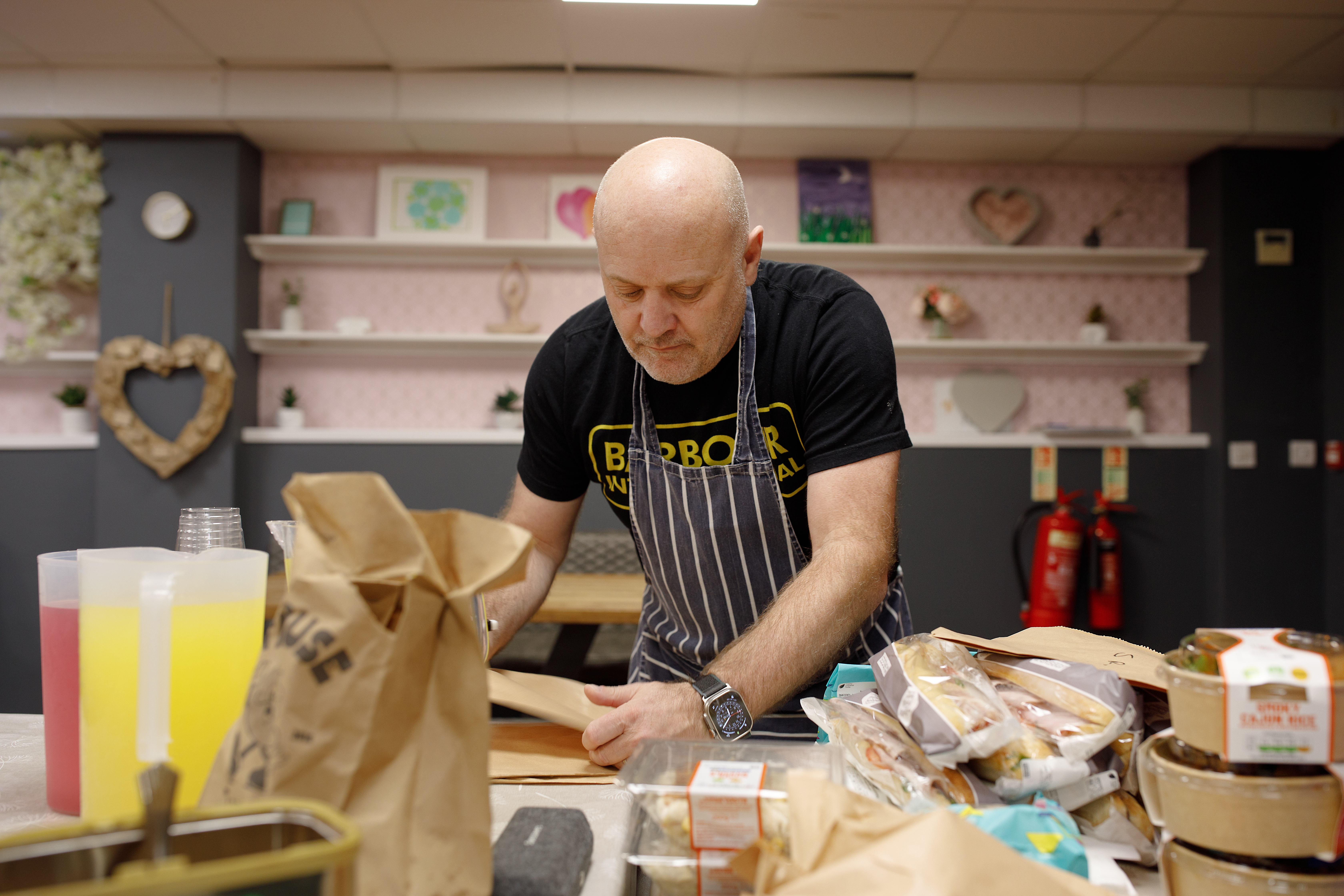 A foodbank volunteer wearing an apron preparing food at a counter covered with paper bags, packaged items, and drinks.