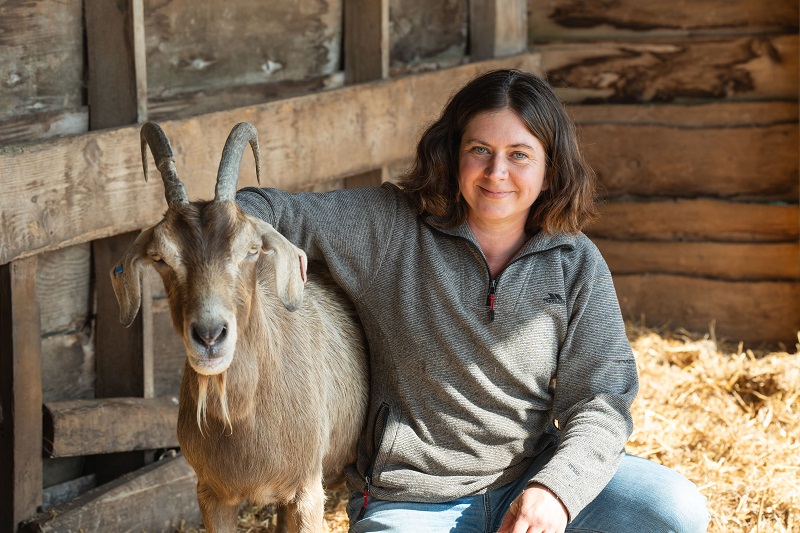 Goat standing outdoors on a smallholding with a female with her arm around the goat.