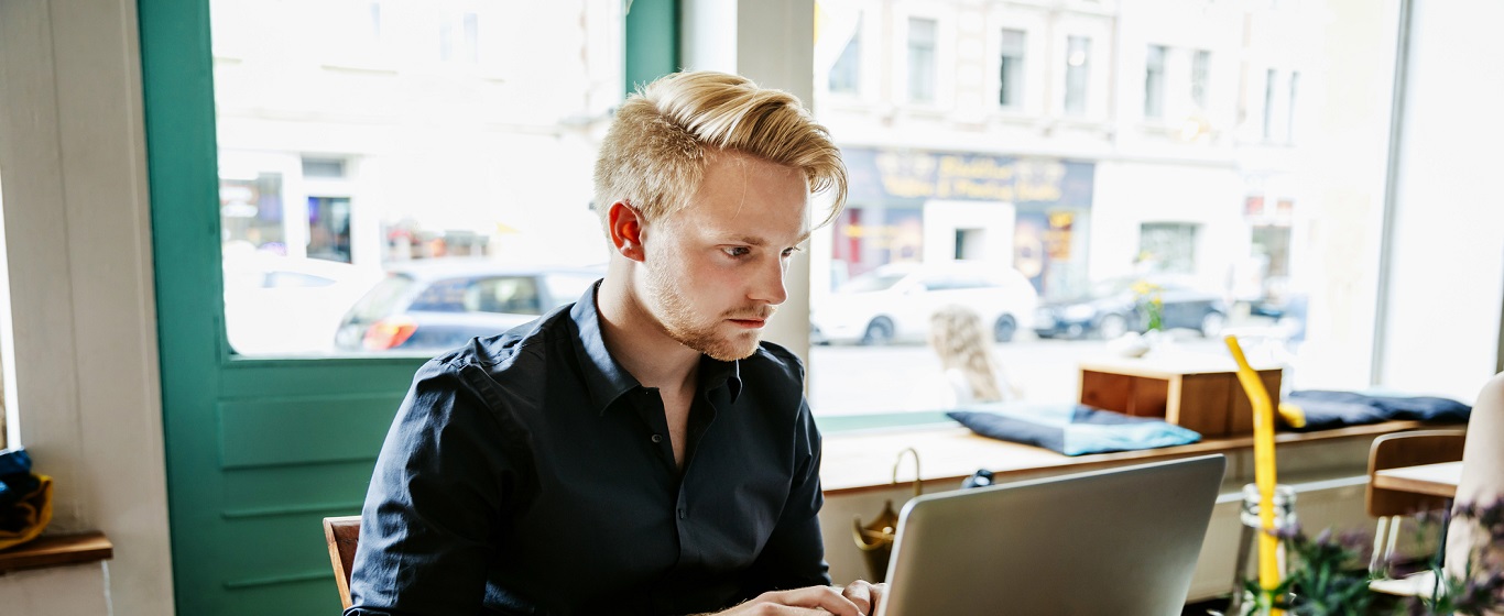 Person working on a laptop at a table inside a café, with large windows showing parked cars and buildings on the street outside.