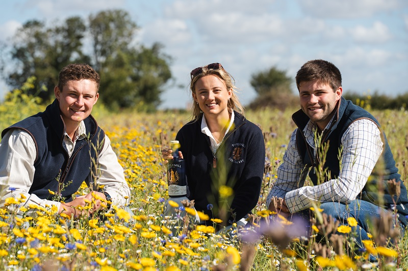 Tipplemill Gin owners, 2 men and 1 woman in the middle, crouching in field of yellow and white daisies