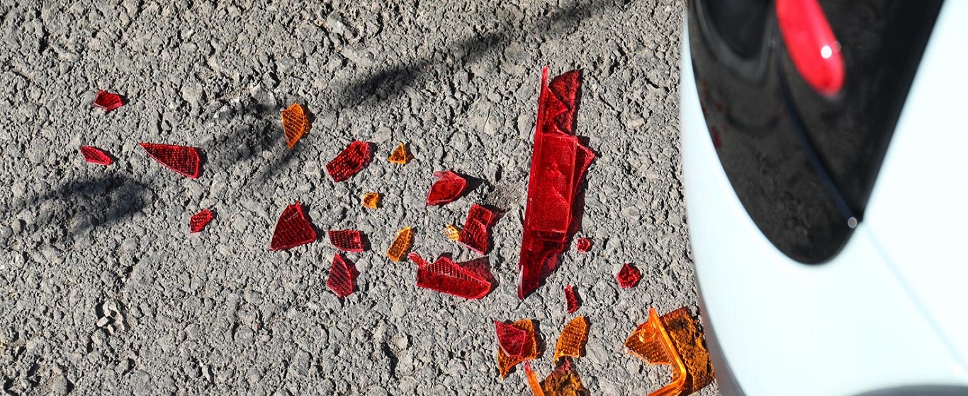 Shattered red and amber car light pieces scattered on rough tarmac beside the rear corner of a white vehicle.