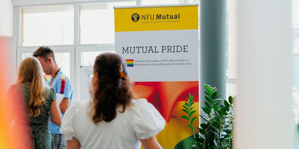 NFU Mutual “Mutual Pride” display banner in an office setting, with colleagues standing nearby and rainbow colours featured on the sign.