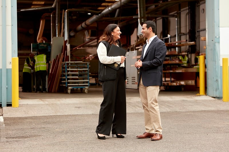 Two people standing and talking outside the entrance of an industrial warehouse, with workers in high‑visibility clothing and equipment visible inside the building.