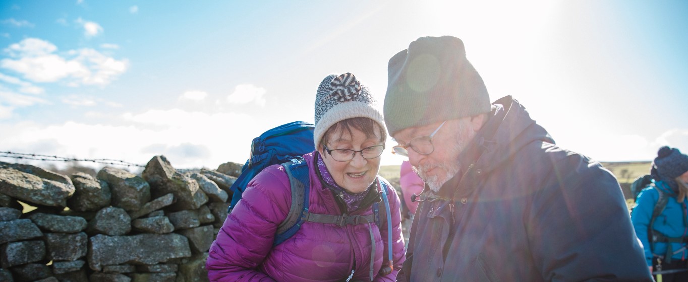 Two hikers in winter clothing standing beside a stone wall, looking down at something together in bright sunlight.