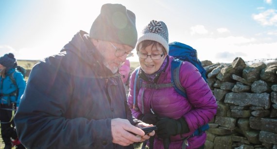 Two hikers in winter clothing standing beside a stone wall, looking down at something together in bright sunlight.