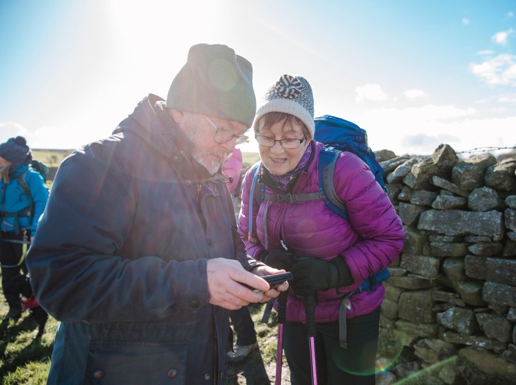 Two hikers in winter clothing standing beside a stone wall, looking down at something together in bright sunlight.