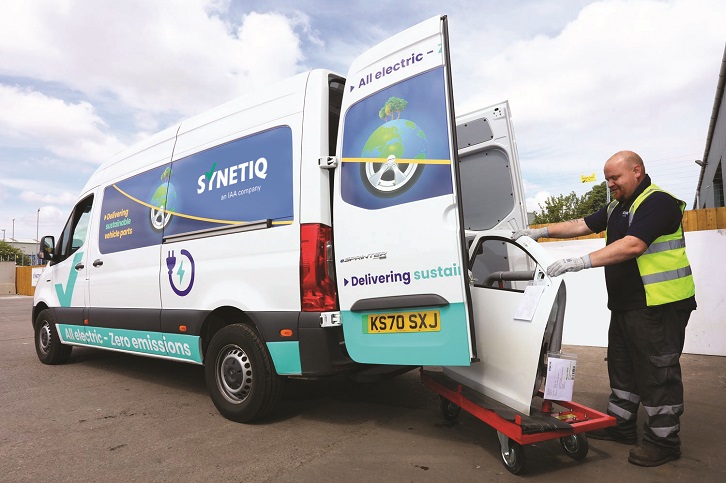 Worker wearing a high‑visibility vest unloads a vehicle part from a trolley into the back of a white SYNETIQ all‑electric van branded with sustainability messaging.