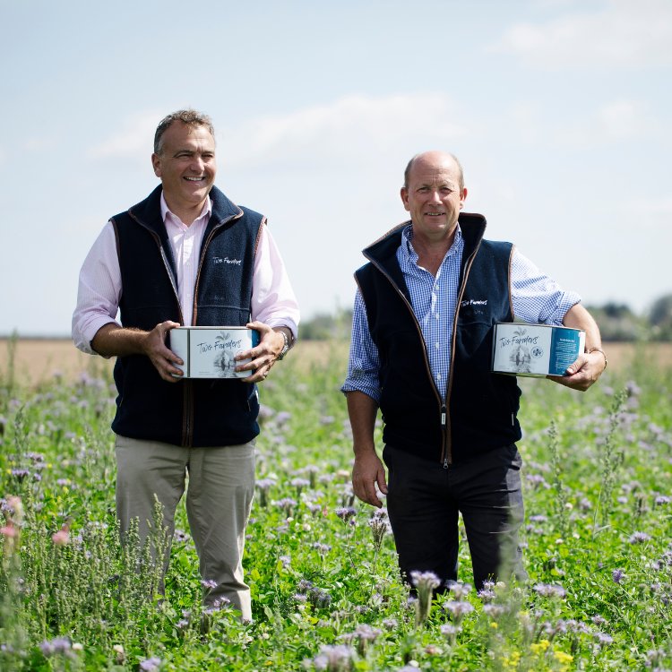 Two farmers standing in a flowering field holding branded product boxes, smiling outdoors under a clear sky.