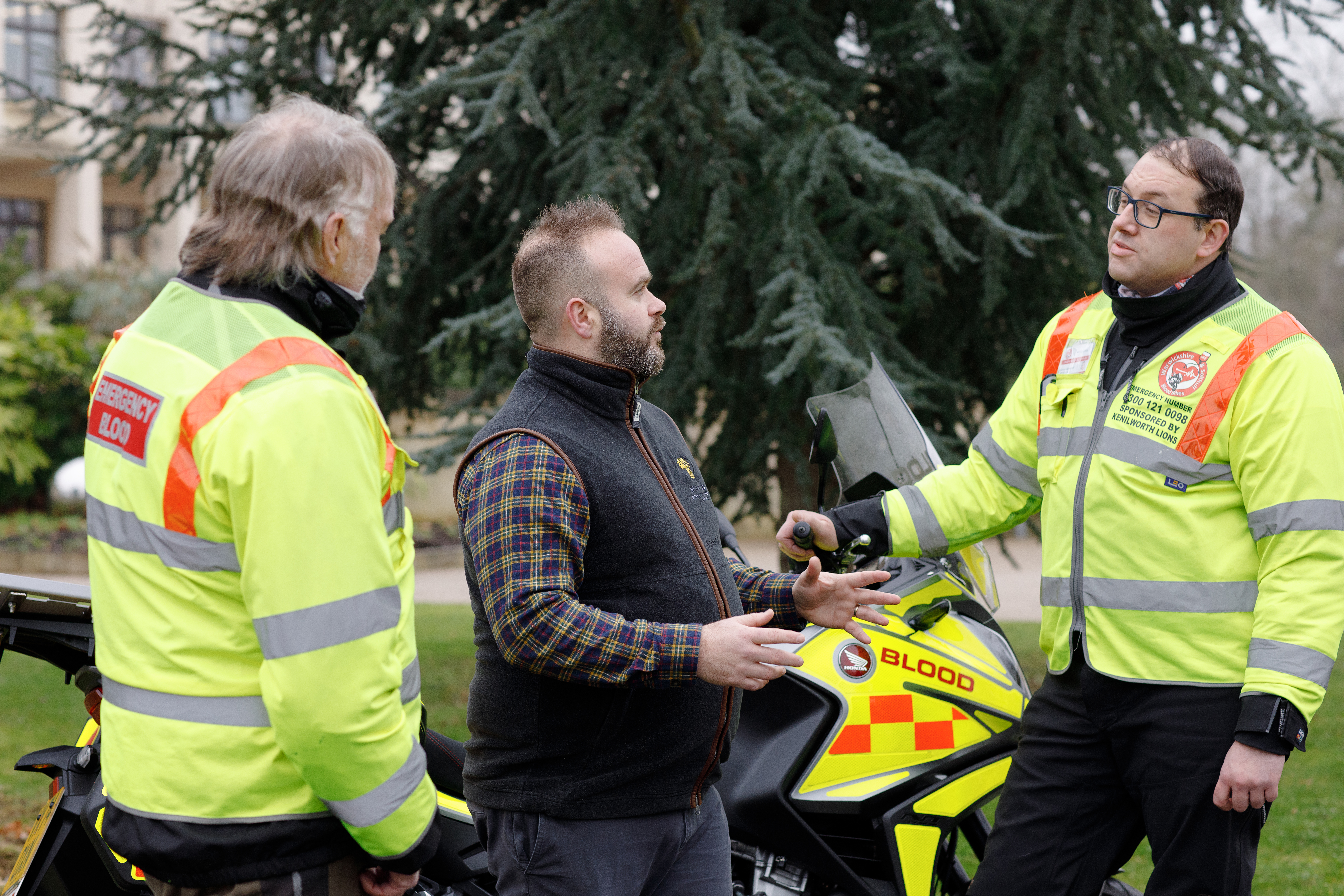 Two emergency responders in high‑visibility jackets talking with a man beside a marked medical motorcycle.