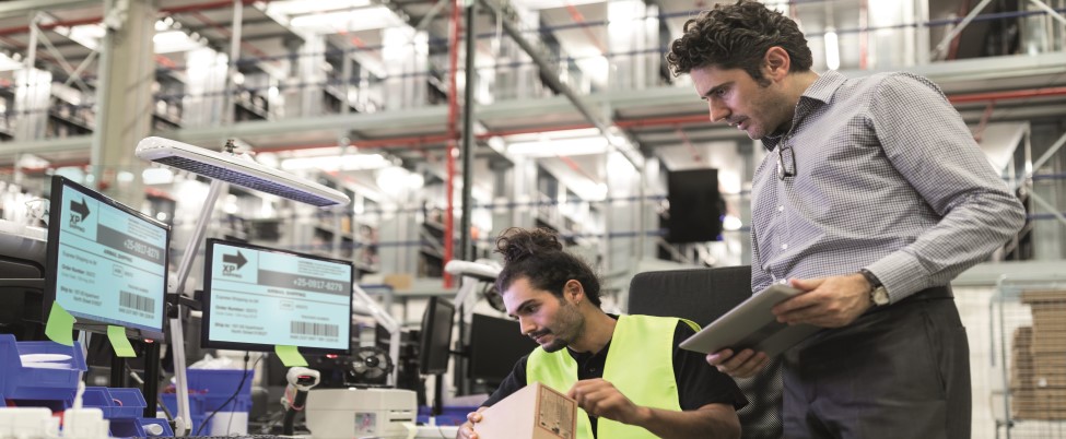 Two warehouse employees reviewing a parcel and shipping details at a workstation, with computer screens and shelving visible in a busy distribution centre.