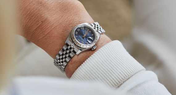 A person wearing a luxury silver wristwatch with a blue dial sits with their arm resting on their lap.