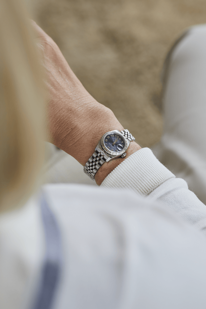 A person wearing a luxury silver wristwatch with a blue dial sits with their arm resting on their lap.
