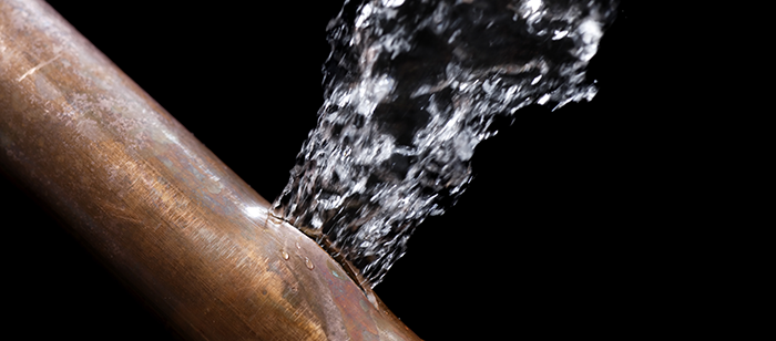 Water spraying forcefully from a crack in a copper pipe against a dark background.
