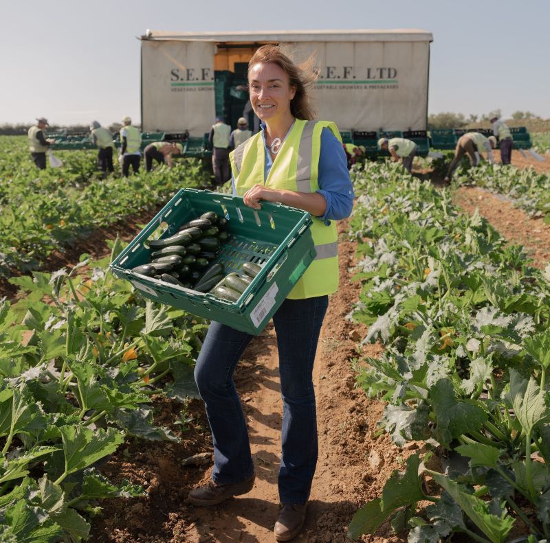 Woman holding a crate of vegetables in farmland, in front of other workers
