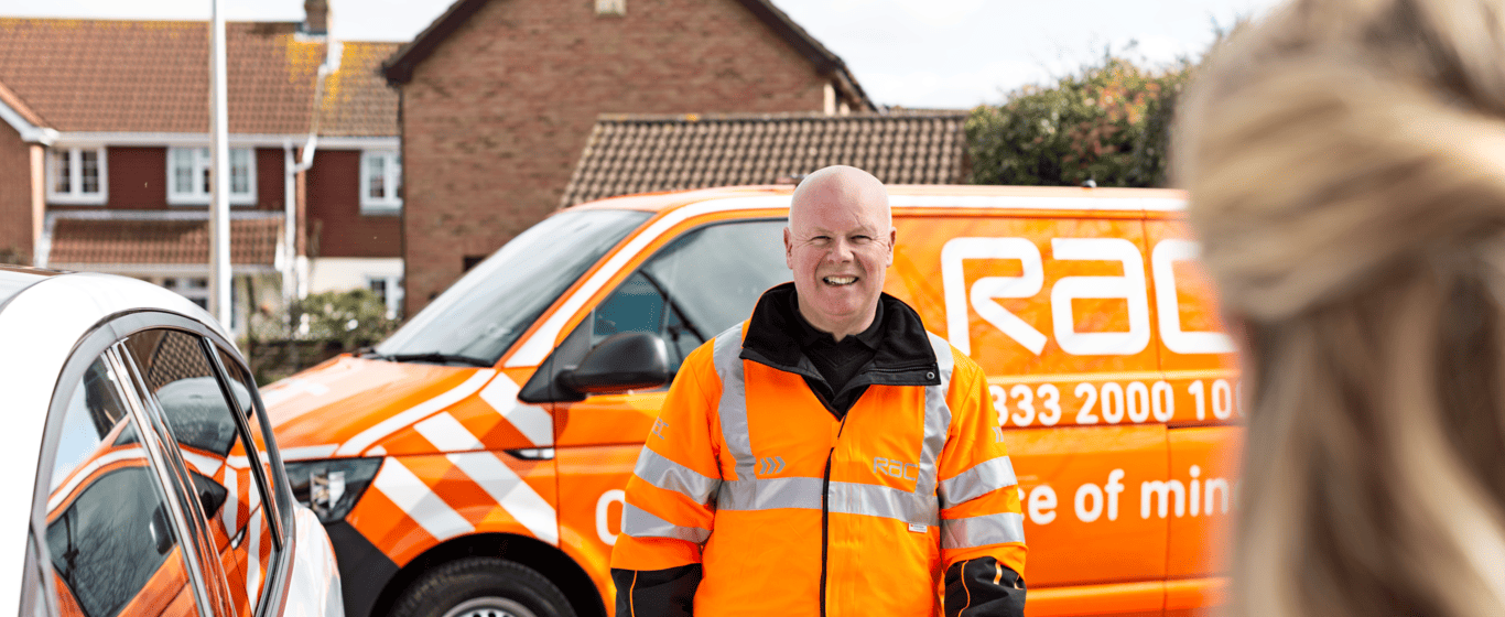Roadside assistance technician in high-visibility orange jacket standing beside a car, with a branded service van parked on a residential street in the background.
